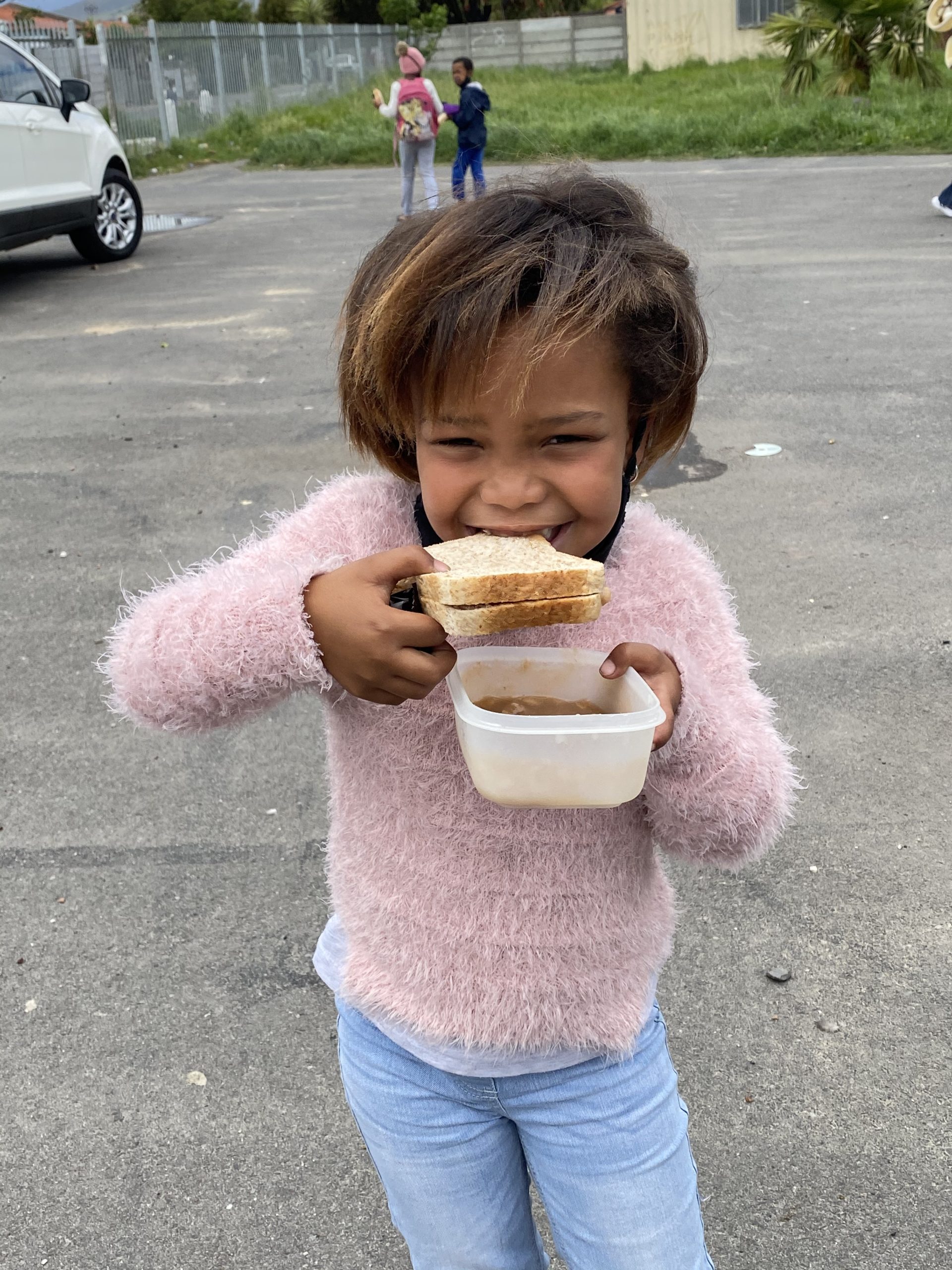 Young girl in pink sweater and jeans smiles as she eats a donated sandwich and holds a bowl of soup