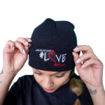 woman standing with her hands on a stylish branded black beanie from the ladles of love merchandise collection